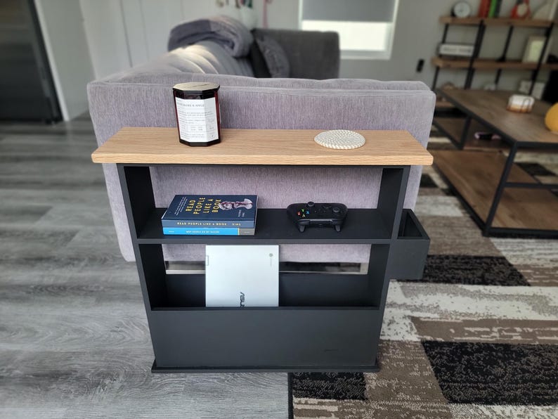 May include: A black and wood side table with two shelves and a small storage compartment. The top shelf holds a candle and coaster. The middle shelf has a book and a game controller. The bottom shelf has a white tablet.