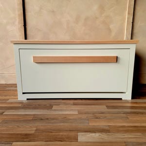 May include: A white storage chest with a light wood top and a horizontal wood handle. The chest is rectangular and sits on a wooden floor. The background is a neutral color.
