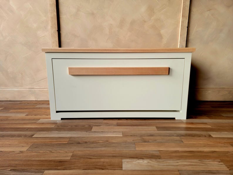 May include: A white storage chest with a light wood top and a horizontal wood handle. The chest is rectangular and sits on a wooden floor. The background is a neutral color.