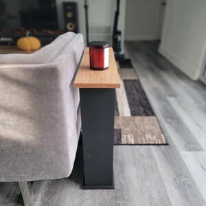 May include: A small side table with a natural wood top and a dark gray base. A red candle in a glass jar with a black lid sits on the table. The table is next to a gray sofa.