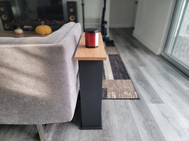 May include: A small side table with a natural wood top and a dark gray base. A red candle in a glass jar with a black lid sits on the table. The table is next to a gray sofa.