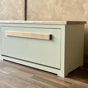 May include: A sage green wooden storage cabinet with a light wood top and handle. The cabinet is rectangular with a single drawer and sits on a wooden floor. The handle is a light-colored wood.