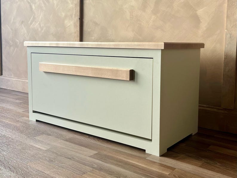 May include: A sage green wooden storage cabinet with a light wood top and handle. The cabinet is rectangular with a single drawer and sits on a wooden floor. The handle is a light-colored wood.