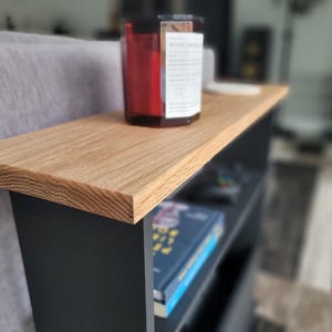 May include: A small, modern side table with a light oak top and black base. A red candle sits on the table's surface. The table has a shelf below for storage, with a book visible. The table is 24 inches long.