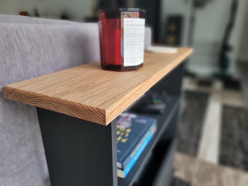 May include: A small, modern side table with a light oak top and black base. A red candle sits on the table's surface. The table has a shelf below for storage, with a book visible. The table is 24 inches long.