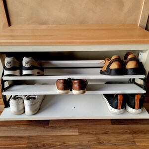 May include: A white and wood shoe storage bench with three shelves, holding various pairs of shoes. The bench has a light wood top and white frame. Shoes include sneakers, loafers, and casual shoes.