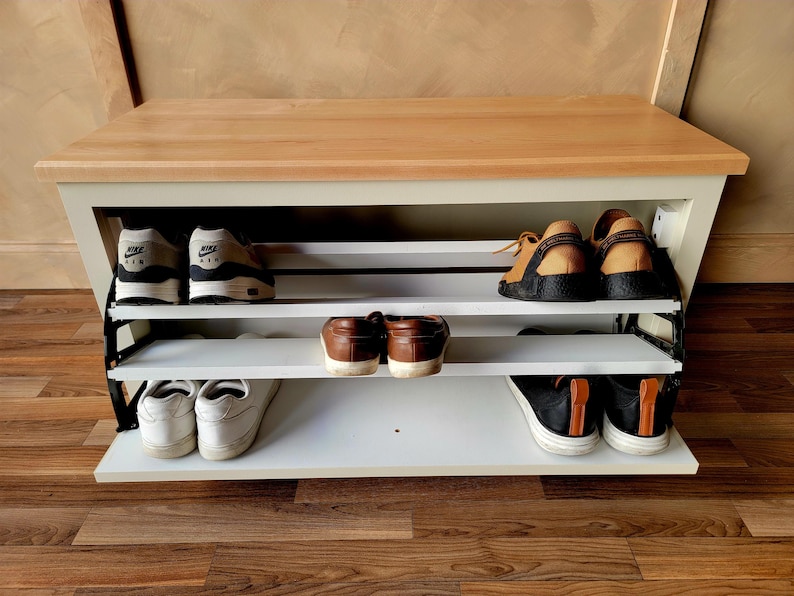 May include: A white and wood shoe storage bench with three shelves, holding various pairs of shoes. The bench has a light wood top and white frame. Shoes include sneakers, loafers, and casual shoes.