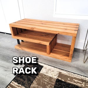 Può includere: Uno scarpiera in legno con due ripiani e una superficie superiore. La scarpiera è realizzata in legno naturale con una finitura marrone chiaro. Il testo "SHOE RACK" è stampato sul pavimento davanti alla scarpiera.
