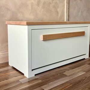 May include: A white cabinet with a light wood top and a horizontal wood handle. The cabinet is rectangular and sits on a wooden floor. The handle is a light brown color.