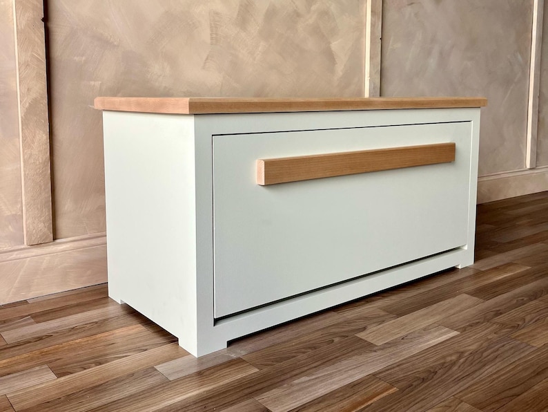 May include: A white cabinet with a light wood top and a horizontal wood handle. The cabinet is rectangular and sits on a wooden floor. The handle is a light brown color.