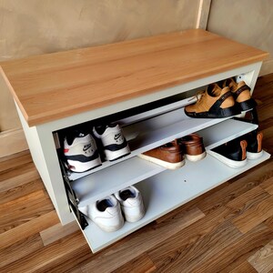 May include: A white and wood shoe storage bench with three shelves, holding various pairs of shoes. The bench has a light wood top and a white base. Shoes include sneakers and leather shoes.