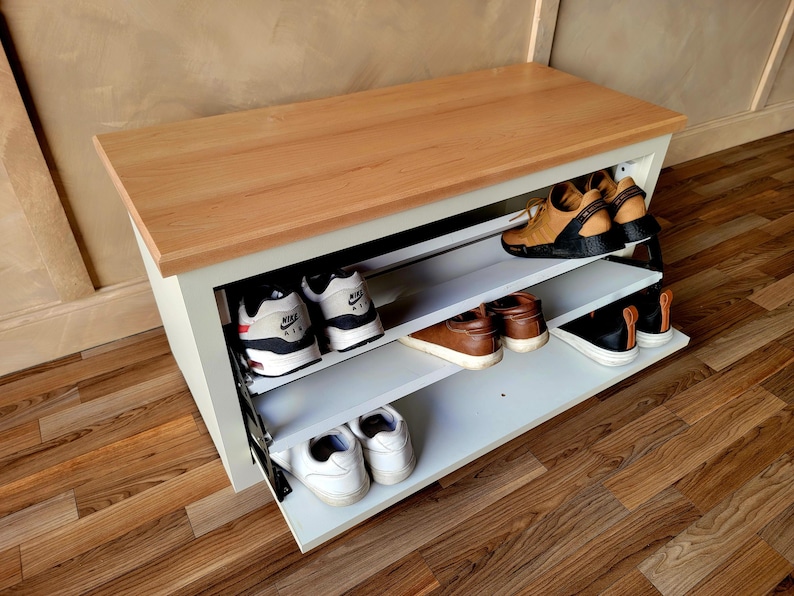 May include: A white and wood shoe storage bench with three shelves, holding various pairs of shoes. The bench has a light wood top and a white base. Shoes include sneakers and leather shoes.