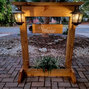 May include: A wooden welcome sign with two lanterns and a planter box. The sign reads "Welcome to our home" in white script. The structure is made of stained wood and has a rustic design. The lanterns are black and have a warm glow.