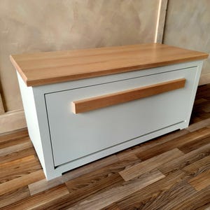 May include: A white storage bench with a light wood top and a horizontal wood handle. The bench is rectangular and sits on a wooden floor. The top is a light brown color.