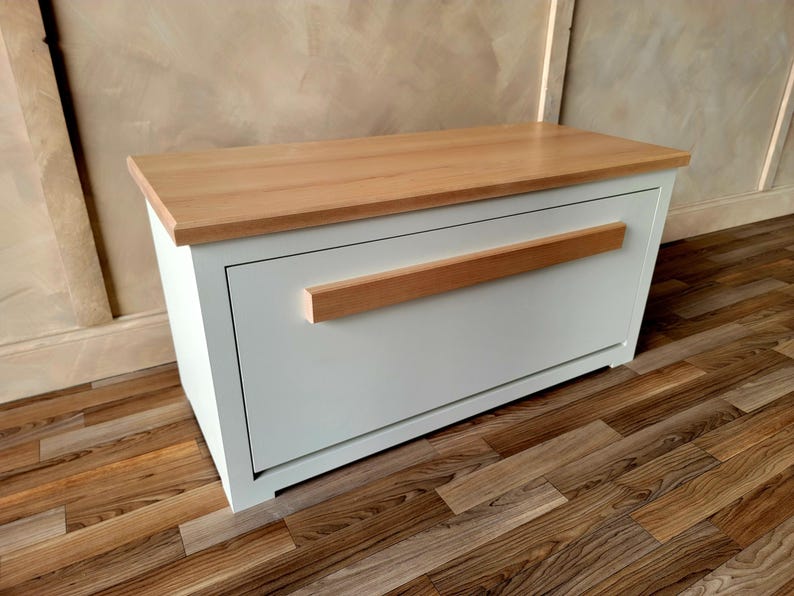 May include: A white storage bench with a light wood top and a horizontal wood handle. The bench is rectangular and sits on a wooden floor. The top is a light brown color.