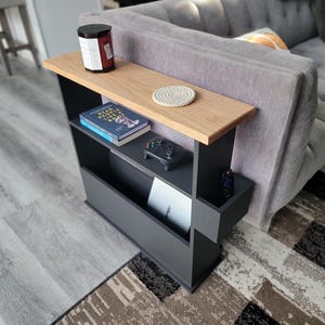 May include: A black and wood-toned side table with a shelf and magazine rack. The table has a light wood top, a shelf with books and a game controller, and a lower magazine rack. A candle and coaster sit on the top.