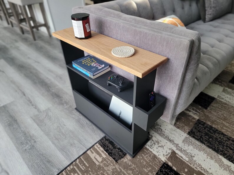 May include: A black and wood-toned side table with a shelf and magazine rack. The table has a light wood top, a shelf with books and a game controller, and a lower magazine rack. A candle and coaster sit on the top.