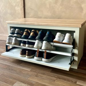 May include: A white and light wood shoe storage bench with two pull-out shelves. The shelves are filled with various shoes, including sneakers and loafers. The bench is set against a neutral-colored wall and wooden floor.
