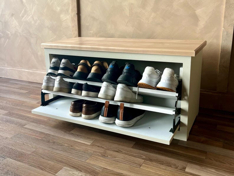 May include: A white and light wood shoe storage bench with two pull-out shelves. The shelves are filled with various shoes, including sneakers and loafers. The bench is set against a neutral-colored wall and wooden floor.