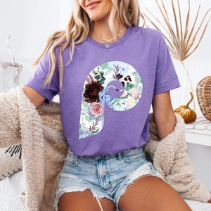 Könnte beinhalten: Ein lila T-Shirt mit einem floralen "P"-Design. Das Blumenmuster enthält bordeauxfarbene, blaue und rosa Blumen mit grünen Blättern. Das Shirt wird mit Jeansshorts getragen.