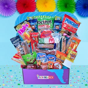 May include: A purple Pachanga Box overflowing with a variety of Mexican snacks and candies. The box contains chips, cookies, peanuts, and candies, with colorful packaging. The background features paper fans in purple, red, yellow, and green.