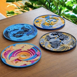 Puede incluir: Cuatro posavasos redondos y coloridos con diseños intrincados. Cada posavasos presenta un patrón único en tonos de azul, amarillo, rosa y blanco. Los posavasos están hechos de papel en capas y están sobre una superficie de madera.