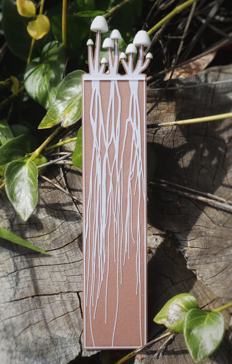 This bookmark has an earthy brown background with delicate white lines resembling intricate fungal root systems or mycelium stretching downward. Mushrooms appear to be growing from the top of the bookmark.