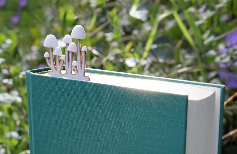 A close-up of a hardcover book with a textured teal cover, placed outdoors in a lush green environment with blurred foliage and purple flowers in the background. A uniquely designed mushgroom bookmark is inserted between its pages. The bookmark is made of a light-wood and white colored pla material, and features a precise silhouette of a small cluster of mushrooms with white caps. The mushrooms appear to be growing from the top of the book, creating a whimsical, nature-inspired aesthetic.