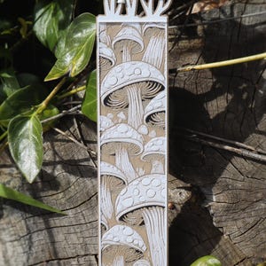This bookmark features an artistic depiction of mushrooms growing in layers, with white outlines and shading against a brown background, creating depth. Mushrooms appear to be growing from the top of the bookmark.