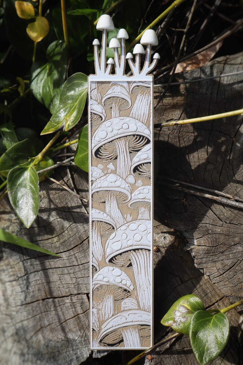This bookmark features an artistic depiction of mushrooms growing in layers, with white outlines and shading against a brown background, creating depth. Mushrooms appear to be growing from the top of the bookmark.