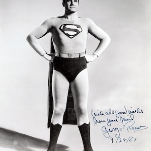 May include: A black and white photograph of a figure in a superhero costume featuring a chest emblem with an S, a cape, a belt, dark trunks, and tall boots. The image includes a handwritten inscription: With all good wishes from your friend George Reeves 9/24/57.