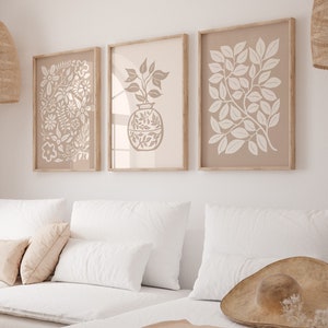 Pode incluir: Um sofá seccional branco com uma manta de lã bege e um chapéu de palha marrom no assento. Três impressões emolduradas com desenhos florais brancos pendurados na parede atrás do sofá.