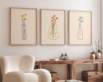 Op de afbeelding: Drie minimalistische line art prints met verschillende bloemstukken in vazen. De prints zijn ingelijst in licht hout en hangen aan een witte muur boven een houten tafel.
