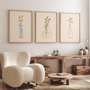Puede incluir: Tres impresiones minimalistas de arte lineal con diferentes arreglos florales en jarrones. Las impresiones est&aacute;n enmarcadas en madera clara y colgadas en una pared blanca sobre una mesa de madera.