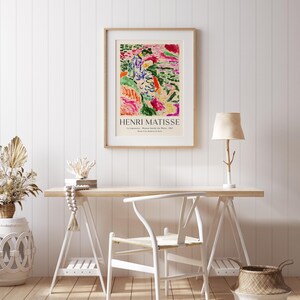 May include: A framed poster with an abstract painting in shades of green, pink, and yellow. The poster is titled "Henri Matisse" and features the text "La Japonaise, Woman Beside the Window, 1951, Museum of Modern Art, Paris". The poster is hanging on a white wall above a wooden desk with a white chair, a lamp, and a basket.