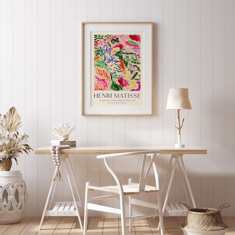 May include: A framed poster with an abstract painting in shades of green, pink, and yellow. The poster is titled "Henri Matisse" and features the text "La Japonaise, Woman Beside the Window, 1951, Museum of Modern Art, Paris". The poster is hanging on a white wall above a wooden desk with a white chair, a lamp, and a basket.