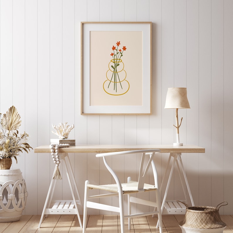 Puede incluir: Una impresi&oacute;n enmarcada de una ilustraci&oacute;n estilizada de un jarr&oacute;n con flores naranjas. El jarr&oacute;n est&aacute; hecho de tres c&iacute;rculos apilados. La impresi&oacute;n est&aacute; colgada en una pared blanca sobre un escritorio de madera con una silla blanca y una l&aacute;mpara.