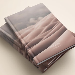 May include: A stack of notebooks with a soft-focus cover design featuring undulating sand dunes in shades of muted pink and beige under a cloudy sky. The front cover displays the text NOTEBOOK and KATE ROSENDALE in a clean white sans-serif font.