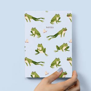 Peut inclure: Un carnet bleu avec un motif répété de grenouilles vertes et de papillons. Le carnet indique "NOTES" sur la couverture.