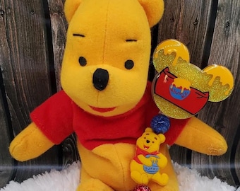 May include: A yellow Winnie the Pooh plush toy with a red shirt is holding a Winnie the Pooh themed retractable badge holder. The badge holder has a blue and red beaded chain with a red glitter charm and a clear plastic Winnie the Pooh charm with the text "Hunny".