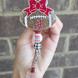 May include: A glittery gold football-shaped badge reel featuring a red glitter bow and a red letter A. Below the football are three beads: one red, one white with the text ROLL TIDE ROLL, and one black and white houndstooth bead with a metal clip.