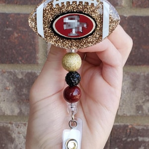 May include: A football-shaped badge reel featuring a gold glitter finish, white laces, and a central red and gold logo. The reel is attached to a vertical strand of three decorative beads in gold, black, and maroon, ending in a clear plastic strap.