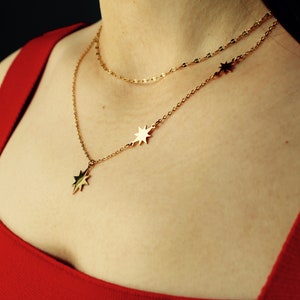 May include: A gold necklace with a star pendant and two smaller star charms. The necklace is layered on a person wearing a red top.