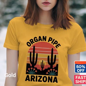 May include: Gold-colored t-shirt featuring a desert-themed graphic. The design includes a sunset, cacti, and the words "ORGAN PIPE ARIZONA" in black. The shirt has short sleeves and a crew neck.