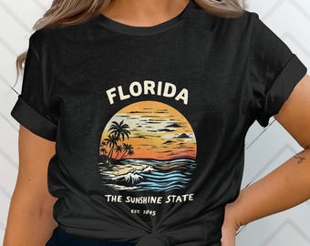 Puede incluir: Camiseta negra con un gráfico de estilo vintage de una puesta de sol sobre una playa con palmeras. El texto "FLORIDA THE SUNSHINE STATE EST. 1845" está impreso en la camiseta.