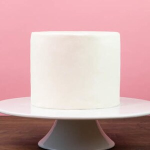 May include: A white cake on a white cake stand. The cake is on a wooden surface and a pink wall is in the background.