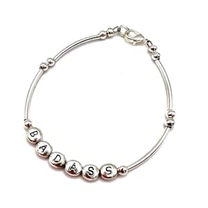 May include: A silver bracelet with the word "BADASS" spelled out in individual round, silver letter beads. The bracelet has a simple, elegant design with a clasp closure and small silver beads along the curved band.