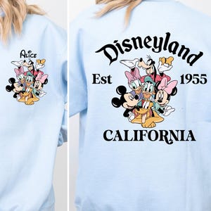 May include: Light blue sweatshirt with a graphic of Mickey Mouse, Minnie Mouse, Donald Duck, Goofy, and Pluto on the front. The back of the sweatshirt has the text 'Disneyland Est 1955 CALIFORNIA'.