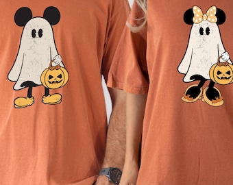 May include: Two orange t-shirts featuring a cartoon ghost with Mickey Mouse ears holding a jack-o'-lantern. One ghost has a bow. The shirts are short-sleeved and have a vintage, distressed look, perfect for Halloween.