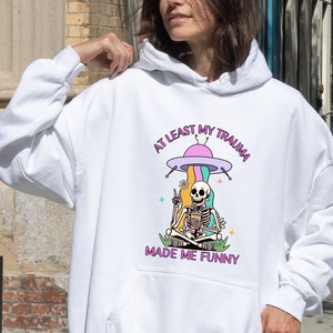 May include: White hoodie with a graphic of a skeleton sitting in a flying saucer with the text "At least my trauma made me funny".